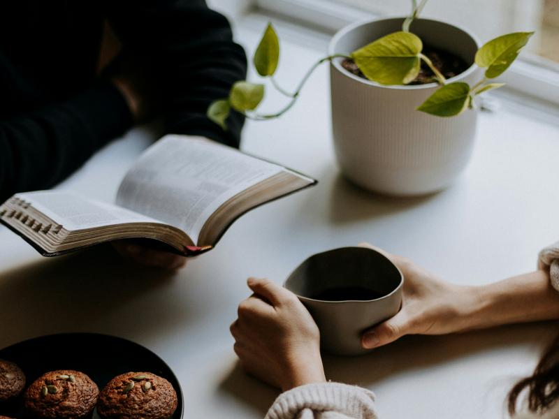 Billeder af en der læser en bog og en anden der sidder med en kop kaffe. Ved bordet er der også kage og en plante.